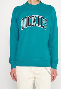 Sweatshirt teal avec col rond à côtes et poignets, arborant "DICKIES" en lettres noires et blanches en gras sur le devant. Coupe standard.