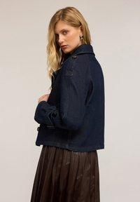 Chaqueta de mezclilla azul marino con detalles de botones, cuello con solapa y corte oversize. Llevada sobre una falda marrón con pliegues, que presenta acentos de costura visibles.