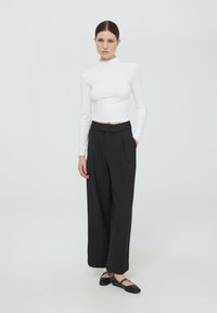 Maglietta bianca a maniche lunghe, aderente, abbinata a pantaloni neri a gamba larga con vita alta. I pantaloni presentano una piega frontale e tasche laterali. Scarpe nere piatte.