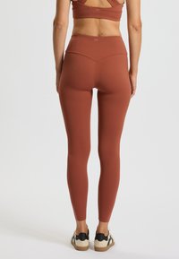 Roodbruin gekleurde legging gemaakt van een rekbaar materiaal, met een hoge tailleband met een dubbelnaadontwerp en een gladde, matte textuur.