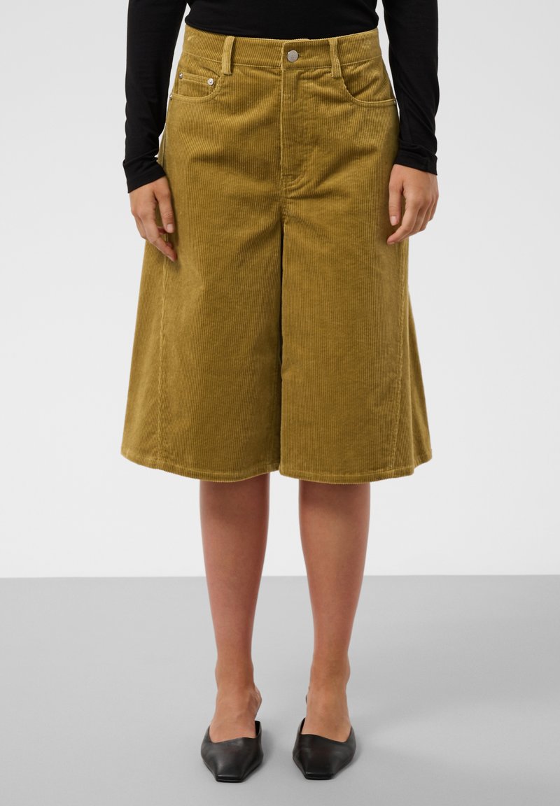 Personne portant un pantalon large en velours côtelé jaune moutarde, un haut noir à manches longues et des ballerines noires pointues, debout sur un sol gris.