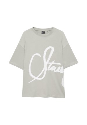 Svetlosivé oversize tričko s krátkymi rukávmi a veľkým bielym kurzívnym písmom čiastočne viditeľným na prednej strane.