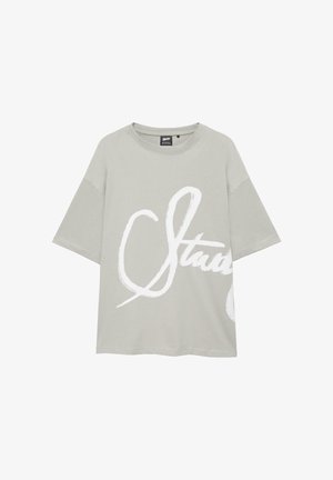 T-shirt oversize grigio chiaro con maniche corte e grande scritta corsiva bianca parzialmente visibile sul davanti.