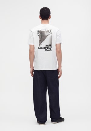 Homme portant un t-shirt blanc avec un logo noir Karl Lagerfeld Jeans dans le dos, pantalon ample foncé et baskets, debout, dos tourné.