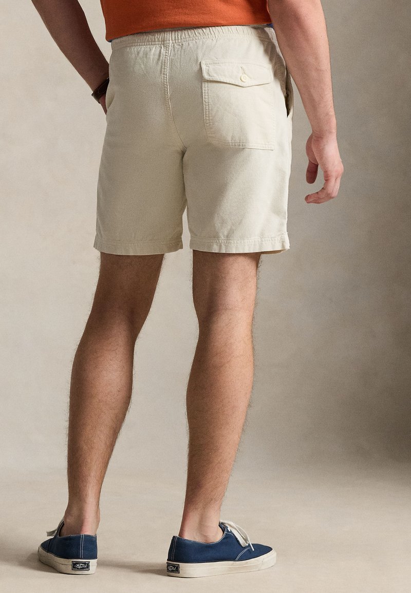 Mann trägt beige Shorts mit Gesäßtasche und blaue Turnschuhe, steht vor neutralem Hintergrund, Hände entspannt an den Seiten.