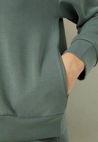 Felpa verde in cotone con orlo e polsini a coste, dotata di una tasca laterale integrata nel design. Texture liscia, vestibilità casual.