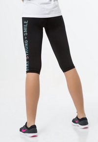Leggings neri cropped con un dettaglio grafico blu e bianco "Be Strong + Smile" sulla gamba sinistra. Abbinati a sneakers nere con dettagli rosa.