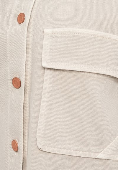 Ελαφρώς μπεζ πουκάμισο με υφασμάτινη υφή, κουμπιά με απόχρωση χαλκού και μπροστινή τσέπη με ραμμένο τελείωμα. Το ύφασμα έχει διακριτικές διαγώνιες γραμμές.