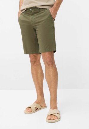 Mann trägt olivgrüne knielange Shorts, beige Sandalen mit überkreuzten Riemen und ein beiges Hemd mit Händen in den Taschen.