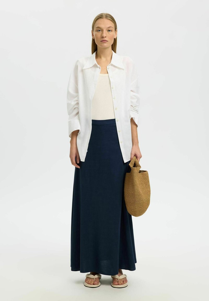 Femme portant une chemise blanche boutonnée, un débardeur beige, une longue jupe bleu marine, des sandales, et tenant un sac à main marron tressé sur un fond uni.