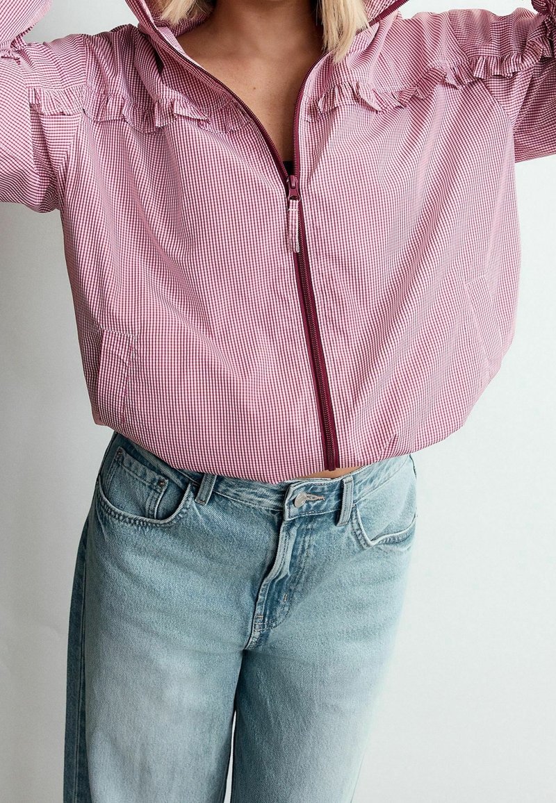 Personne portant une veste zippée à carreaux roses et blancs avec un jean en denim bleu clair, ajustant la capuche de la veste.