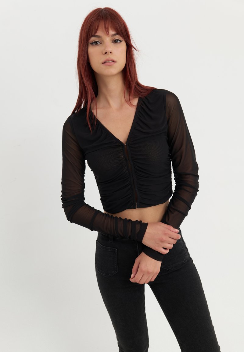 Svart cropped topp med gjennomsiktige lange ermer, rynket design, V-hals og glidelås foran, kombinert med høyde idyll svart jeans.