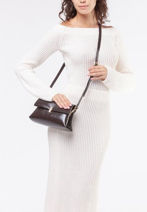 Borsa a tracolla in pelle marrone con chiusura a pattina e hardware dorato. La modella tiene la borsa contro un vestito lavorato a maglia crema.
