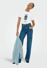 T-shirt bianca con grafica e testo blu, abbinata a jeans blu a vita alta. La modella tiene un blazer azzurro. Tessuto testurizzato e finitura liscia.