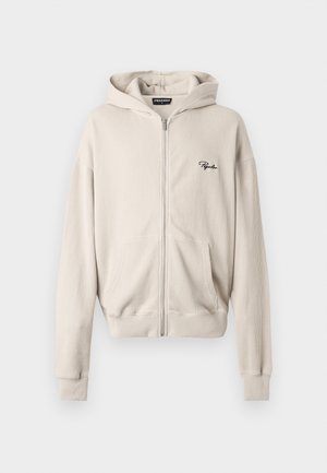 Beige ritsjack in ribbelstof, met een voorzak, zachte textuur en een klein zwart logo op de linkerkant van de borst.