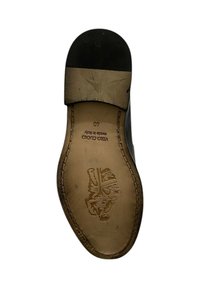 Parte inferiore di una scarpa nera in pelle con tacco piatto, bordi cuciti e logo in rilievo. Presenta una soletta interna in pelle liscia color tan e segni di usura.