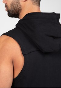 Zwarte mouwloze hoodie met een getailleerde pasvorm, voorzien van een verstelbare capuchon, zachte textuur en nette stikdetails op de schouder.
