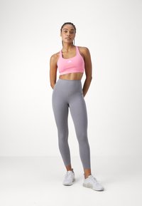 Roza športna modrčka z belim logotipom, v kombinaciji z visokimi sivo-lasičarji. Model ima na sebi bele športne čevlje in stoji na enostavni podlagi.