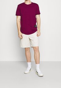 Hombre vestido con una camiseta de manga corta morada oscura, pantalones cortos beige, calcetines blancos y zapatillas blancas con acentos negros, de pie frente a un fondo liso.