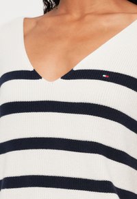 Weißer Strickpullover mit navyblauen horizontalen Streifen, ausgestattet mit einem tiefen V-Ausschnitt und einem kleinen Logo-Patch in Rot, Weiß und Blau auf der Brust.