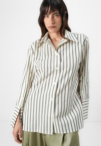 Long-sleeve button-up shirt in cream with black vertical stripes, featuring a classic collar and side slits at the hem for added detail.