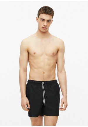 Jeune homme aux cheveux courts et bouclés portant un short de bain noir à cordon, debout devant un fond blanc.