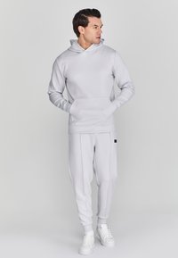 Lichtgrijze hoodie met een Buikzak, gecombineerd met bijpassende tapered sweatpants. De stof lijkt glad met elastische boorden bij de enkels.