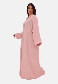 Una donna sta in profilo, indossando un lungo vestito rosa chiaro con maniche ampie e colletto, che mette in risalto la sua vestibilità rilassata e la trama.