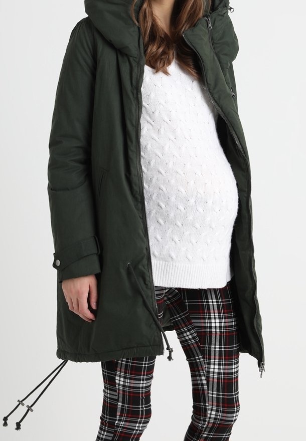 Těhotná žena v tmavě zeleném parku, bílém pleteném svetru a kalhotách s kostičkovaným vzorem v černé, bílé a červené barvě na neutrálním pozadí.