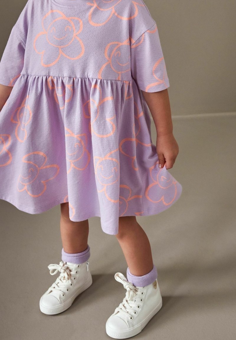 Tout-petit portant une robe lavande avec des imprimés de fleurs souriantes orange, des chaussettes lavande et des baskets montantes blanches, debout sur un sol gris.