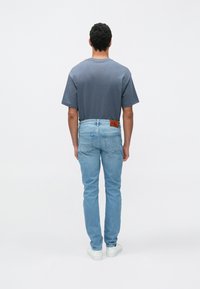 Siva majica s kratkimi rokavi, kombinirana s svetlo modrimi kavbojkami in belimi supergami. Opazna rjava logotipna značka na zadnjem delu kavbojk.