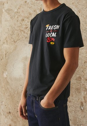 Osoba ubrana w czarną koszulkę z napisem "Fresh & Local 574" w kolorach białym i czerwonym, w ciemnoniebieskich dżinsach, stojąca na tle beżowej teksturowanej ściany.