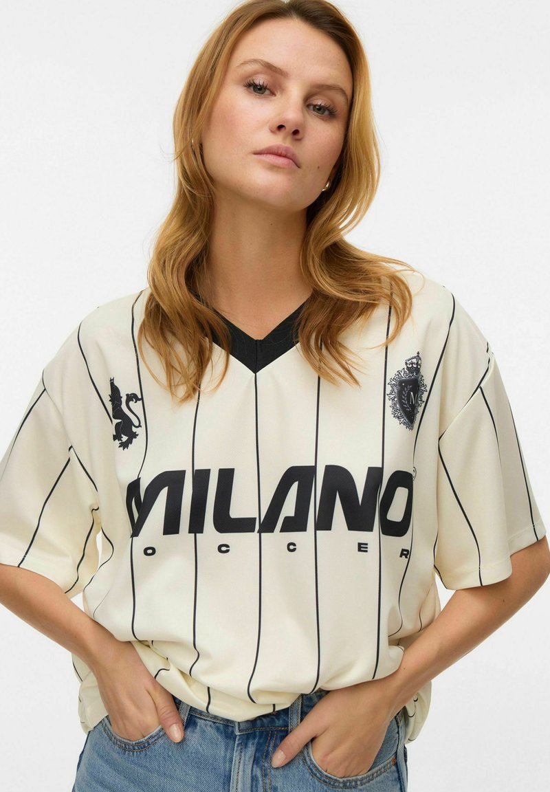 Cremefarbenes Fußballtrikot mit schwarzen vertikalen Streifen, V-Ausschnitt und schwarzer Schrift "MILANO SOCCER". Verfügt über Embleme auf beiden Seiten.