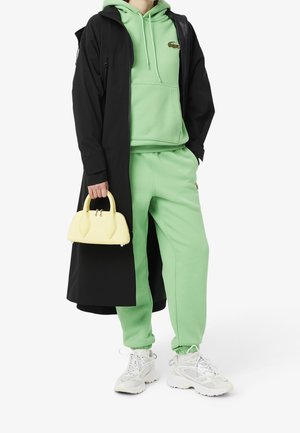 Felpa verde menta e pantaloni della tuta abbinati a un lungo cappotto nero, sneakers bianche e una piccola borsa gialla con manici arrotondati.