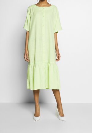 Frau trägt ein locker sitzendes, hellgrünes Midi-Hemdblusenkleid mit kurzen Ärmeln und volantsaum, kombiniert mit weißen High-Heel-Schuhen.