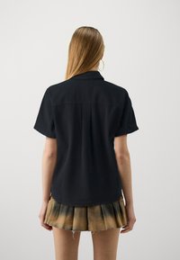 Camisa negra de manga corta con cuello, de corte recto y textura lisa, llevada sobre una falda plisada de tonos beige y marrón.