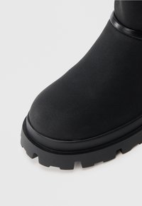 Melnas potītes zābaki no gluda materiāla ar apaļu purngalu, biezu melnu zoli ar dziļiem protektoru rakstiem un smalku tekstūru.