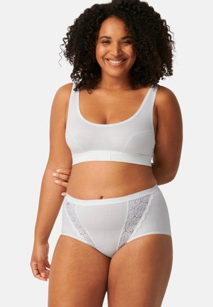 Femme souriante aux cheveux bouclés portant un soutien-gorge en coton blanc et un slip taille haute avec des détails en dentelle, debout devant un fond uni.