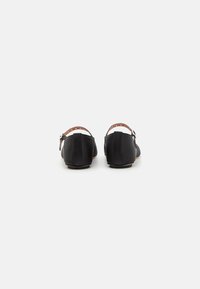 Scarpe nere con punta tonda, texture liscia e tacco basso. Presentano una cinghia marrone regolabile con hardware in metallo e ritagli circolari.