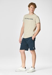 T-shirt di cotone beige con testo scuro blu "U.S. POLO ASSN.", abbinato a pantaloni corti blu navy. Il modello indossa sneakers bianche, con le mani nelle tasche.