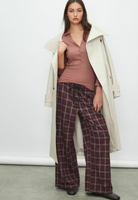 Dugi kremasti trenkč kaput preko mauve henley majice, u kombinaciji sa širokim ljubičastim kariranim hlačama i tamnim cipelama. Glatke teksture, neutralna paleta boja.