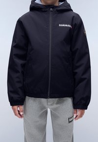 Svart jacka med dragkedja och huva, med en logo på bröstet och elastiska muddar. Matchas med grå mjukisbyxor. Slät textur.