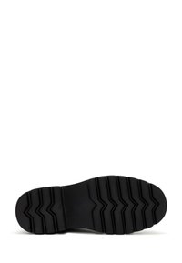 Suela de zapatilla de goma negra con un patrón de banda de rodadura en zigzag, que presenta secciones elevadas para agarre y textura. Área de los dedos suave y redondeada.