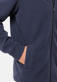Giacca in pile blu navy con superficie testurizzata, chiusura con zip e tasche laterali. Il tessuto appare morbido e caldo, ideale per il clima fresco.