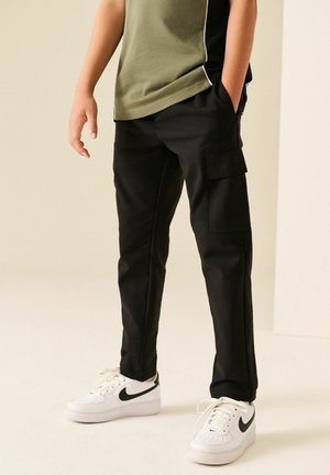 Kind draagt zwarte cargobroek, witte Nike-sneakers en een olijfgroen shirt met handen in de zakken, staand tegen een lichte achtergrond.