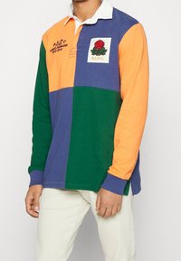 Färgblockerad långärmad polo i orange, lila och grön, med en vit krage. Har en broderad ros och text på bröstet.