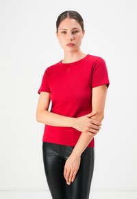 Woman with dark hair in a red short-sleeve top and black leather pants stands against a plain white background, holding her left arm.