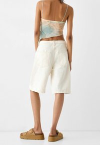 Shorts in denim bianco con vestibilità comoda, dotati di quattro tasche. Il modello indossa una maglia tie-dye e sandali platform marroni.