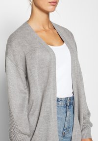 Cardigan lavorato a maglia grigio chiaro con apertura frontale, maniche larghe e un motivo testurizzato. Indossato sopra un top bianco e jeans a vita alta blu chiaro.