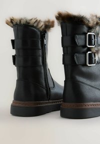 Botas negras con borde de pelo sintético, cierre de cremallera y dos acentos de hebilla. Presentan un material texturizado similar al cuero y una suela de goma resistente.